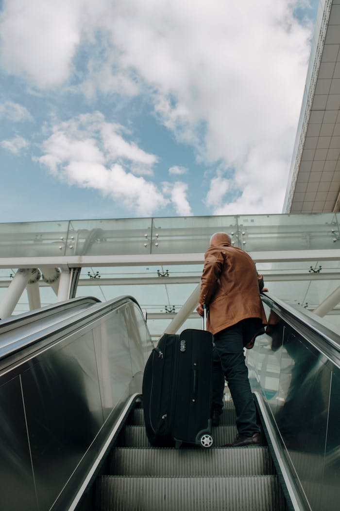 Adult male riding an escalator with luggage in an airport, traveling with bright sky overhead.