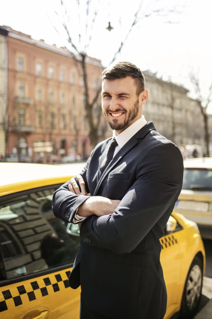 A confident businessman in formal attire stands smiling by a yellow taxi on a sunny day.