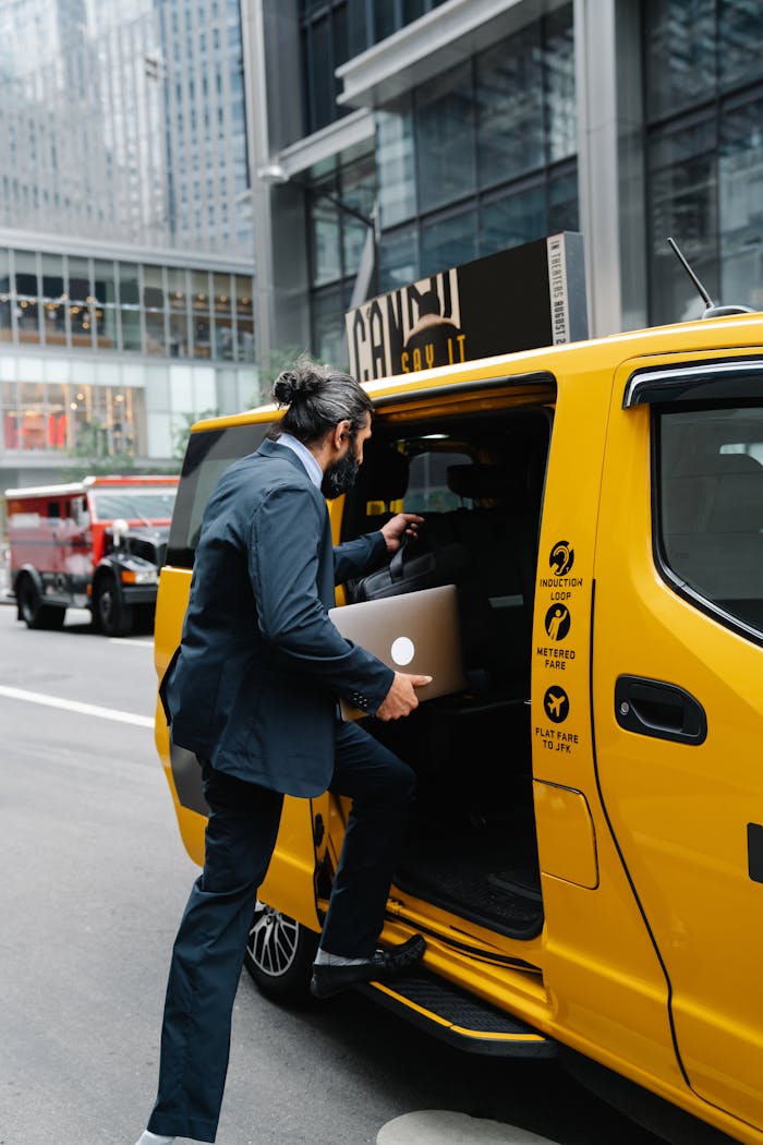 heros-img A businessman in formal attire enters a yellow taxi in a bustling city area, holding a laptop.